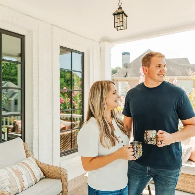 A blonde woman stands next to a blond man on a porch holding a copper mug in front of black Infinity casement windows.