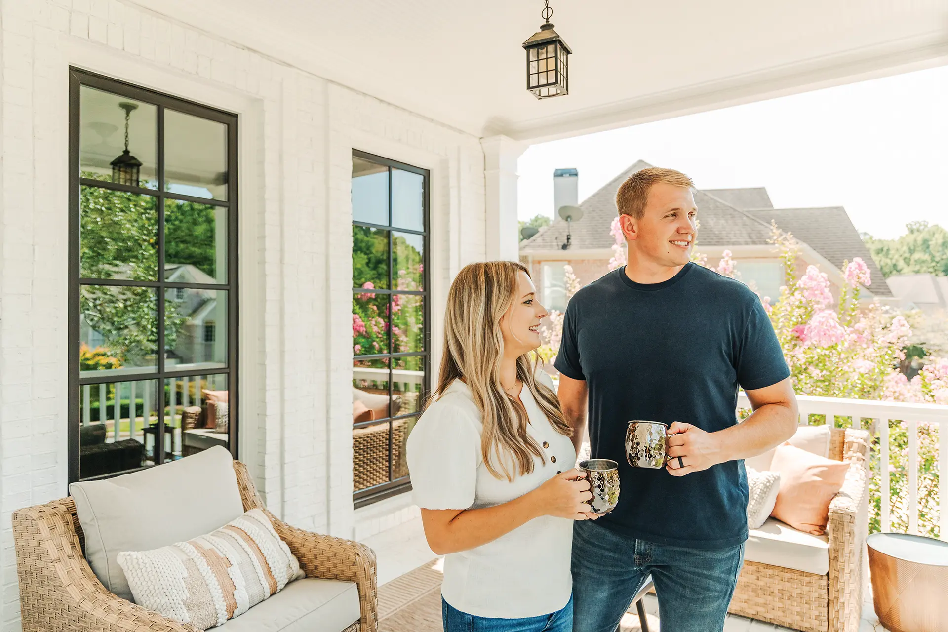 filtersquality100filltransparent (1) A blonde woman stands next to a blond man on a porch holding a copper mug in front of black Infinity casement windows.